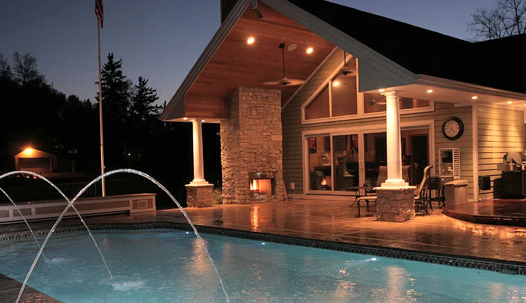 Phoenix pool and spa in Coloma, MI at night, with deck jets arcing in front of the lit porch and stone fireplace