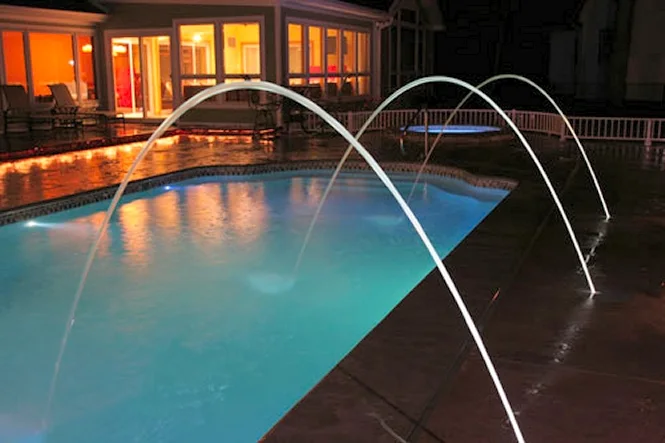 Deck jets arcing across the pool at night in front of the lit house