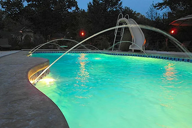 A deck jet arcing into the lit pool at dusk beside the slide