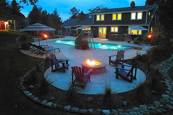 Adirondack chairs around the fire pit at night with the pool lit behind